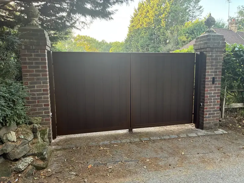 Pair of dark brown solid-panel driveway gates with vertical boarding, closed between two red brick piers topped with stone finials, with a keypad mounted on the right-hand pier