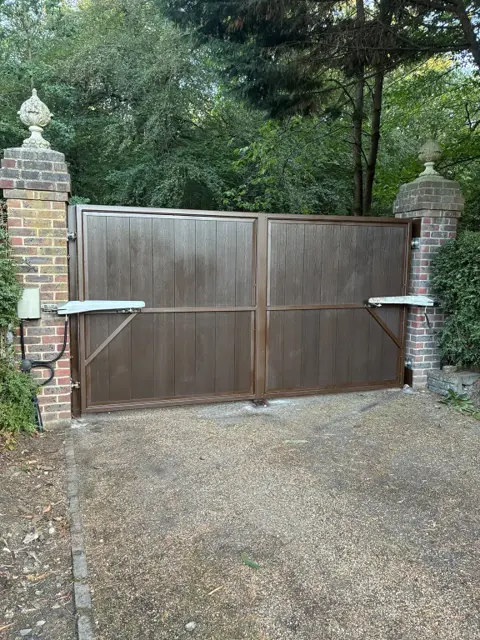 Inside view of closed brown double swing gates between brick piers, each leaf fitted with an articulated-arm gate motor mounted on the pier, with diagonal frame bracing and a gravel driveway in front