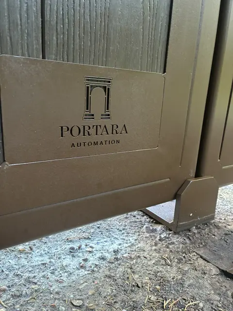 Close-up of a brown metal gate frame with a cut-out Portara Automation logo panel below wood-grain boarding, and a metal gate stop at the base where the two gate leaves meet